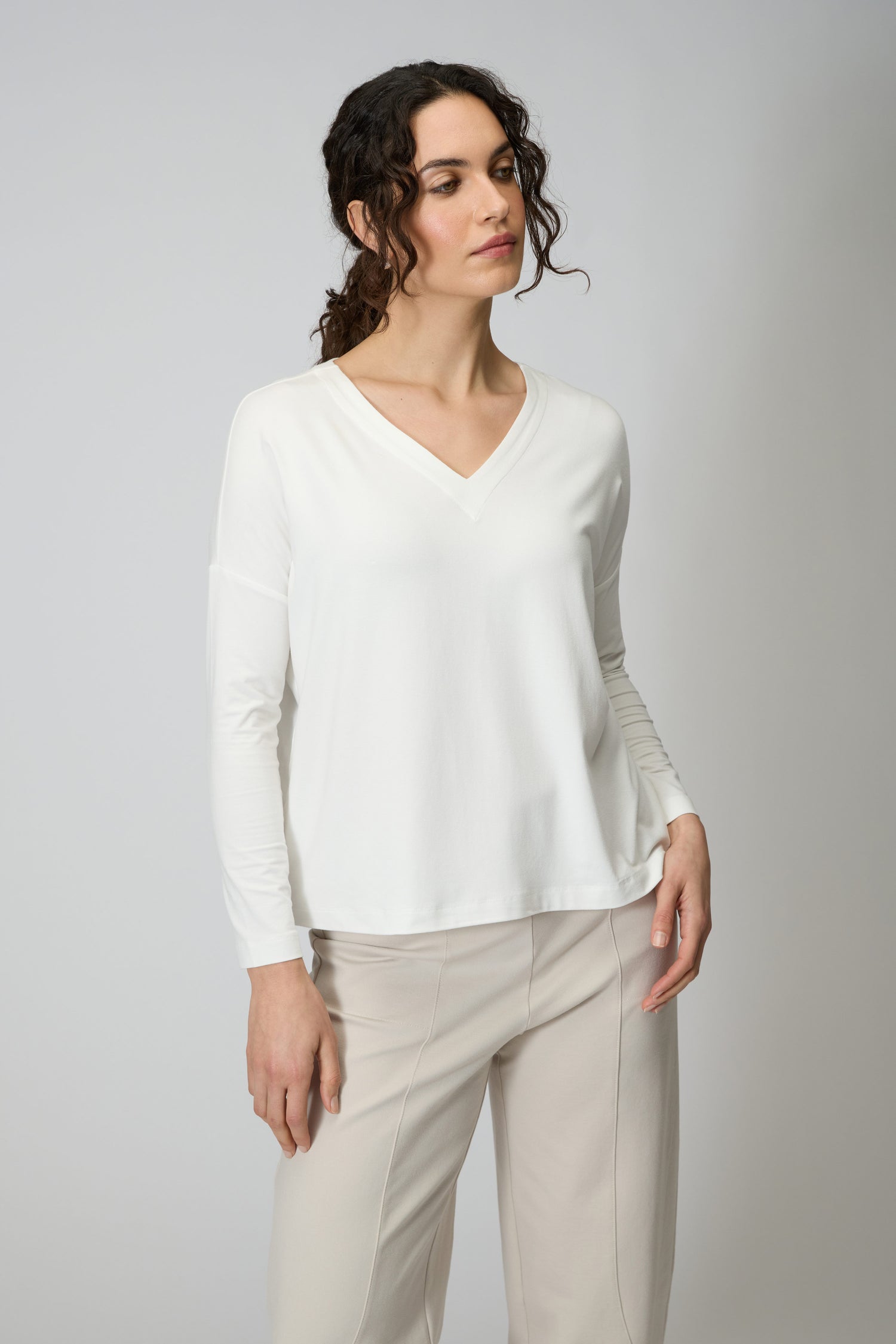 A woman stands against a plain light gray background wearing the Viscose Jersey V Neck Top in white with long sleeves, paired with light beige pants.