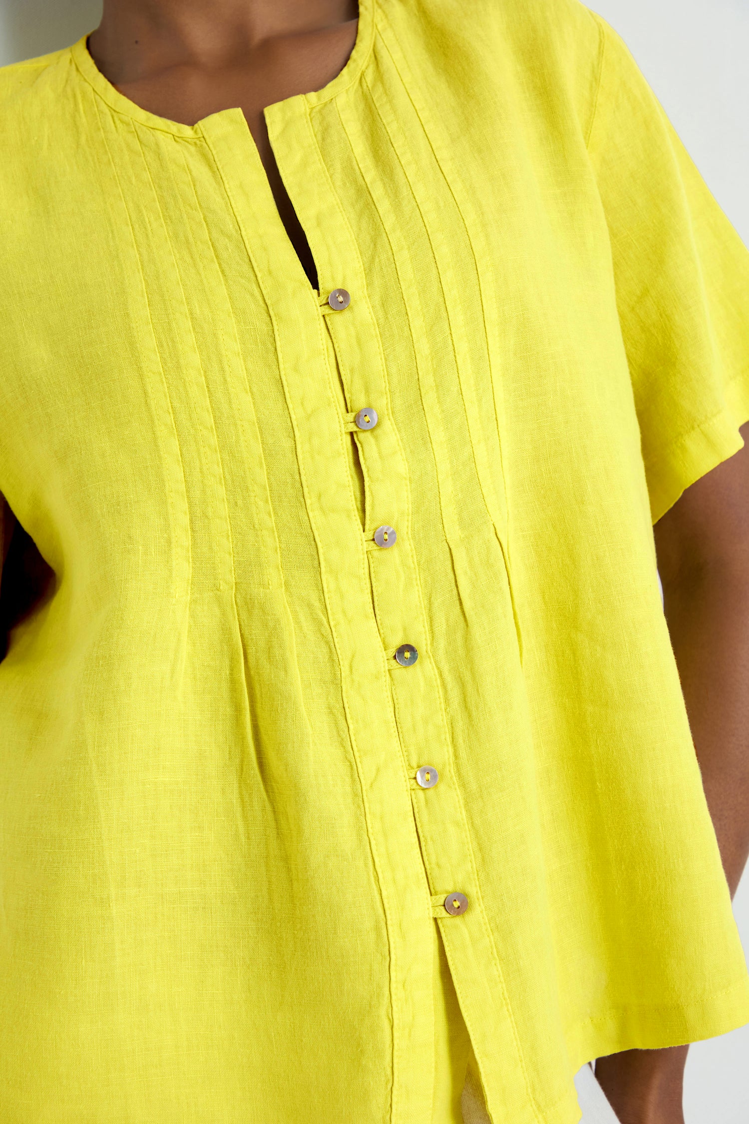 The Linen Pleat Short Sleeve Lemon Shirt features a bright yellow hue, lightweight linen, button-up front, and pleated details—perfect for adding a stylish touch to any summer outfit.