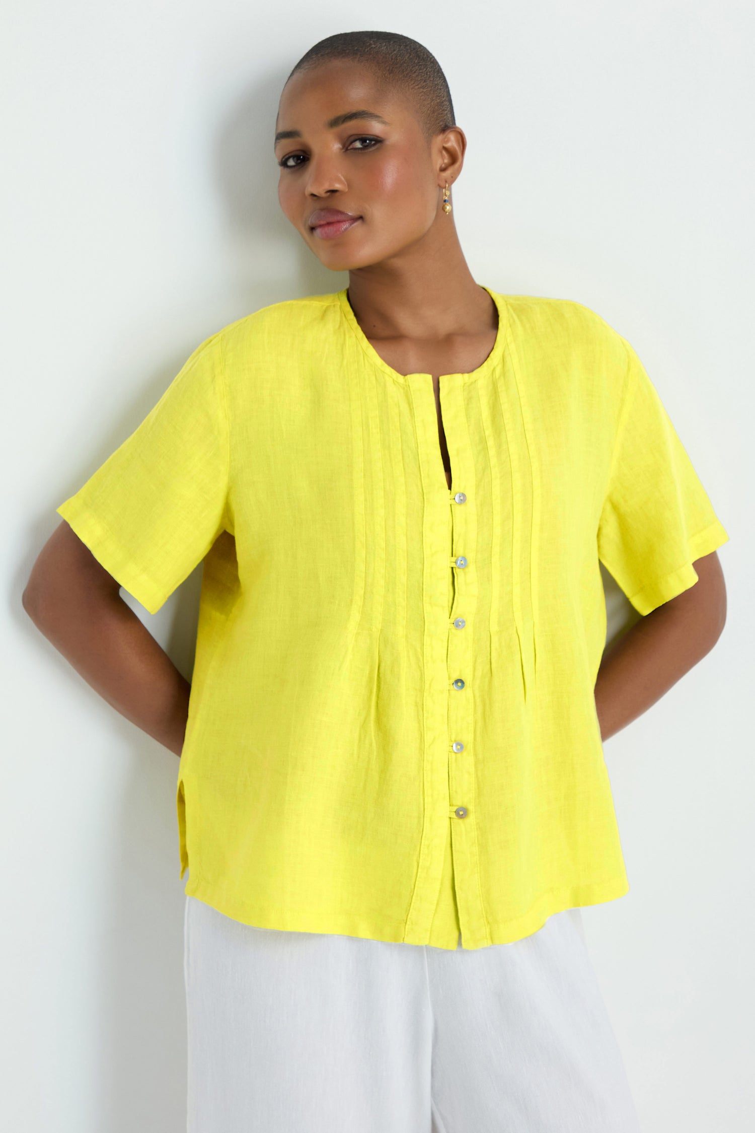 A person stands against a white wall, wearing the Linen Pleat Short Sleeve Lemon Shirt with a button-front design, paired with crisp white pants.
