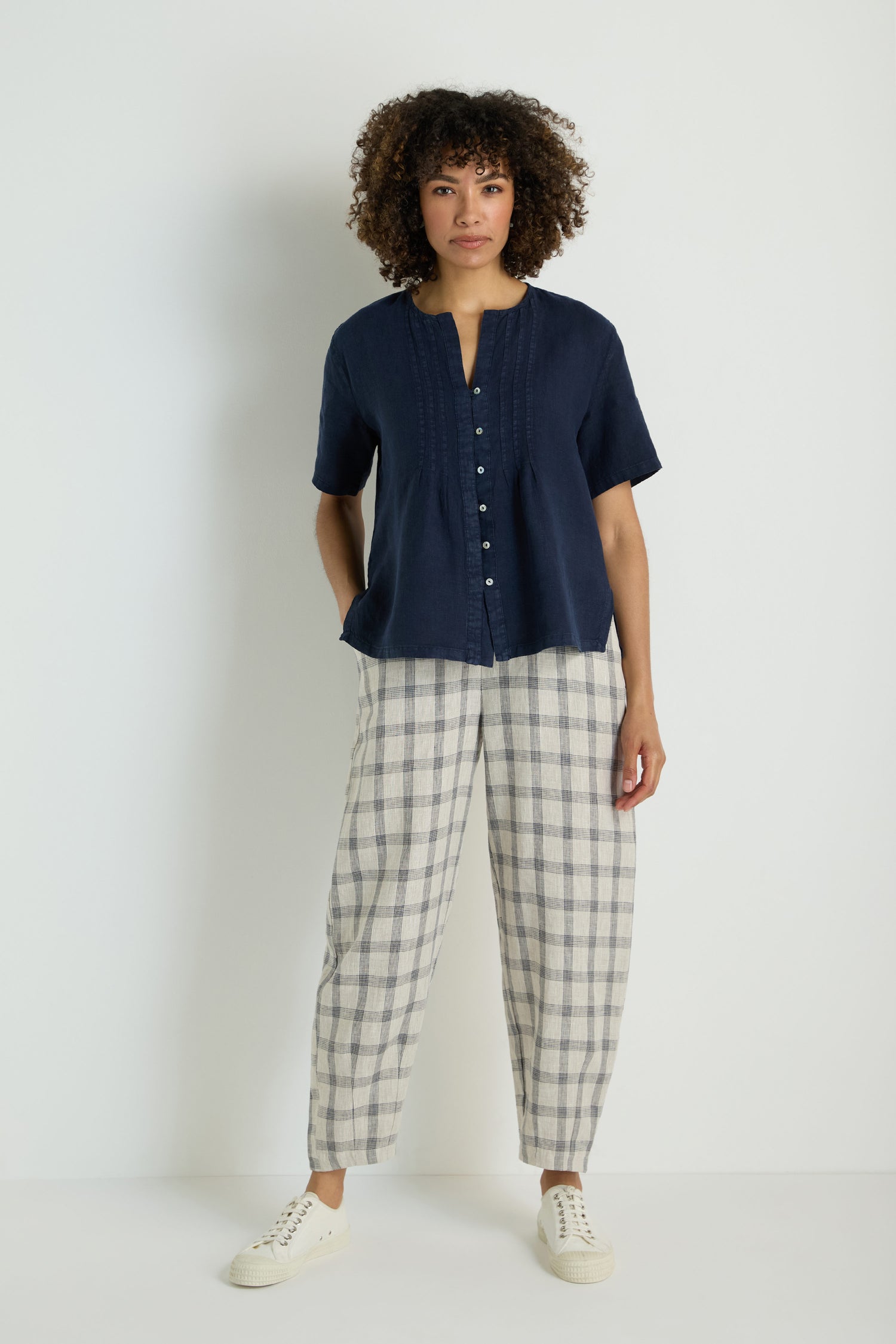 A woman stands against a white wall, wearing the Linen Pleat Short Sleeve Shirt in navy, paired with light gray wide-leg pants and white sneakers.