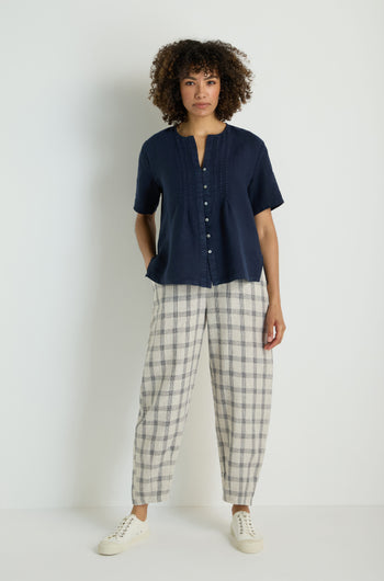 A woman stands against a white wall, wearing the Linen Pleat Short Sleeve Shirt in navy, paired with light gray wide-leg pants and white sneakers.