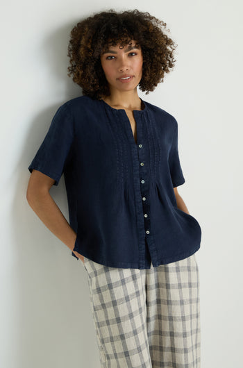 Wearing the Linen Pleat Short Sleeve Shirt in navy blue and light gray pants, a person sits on a light wooden box, looking to the side.