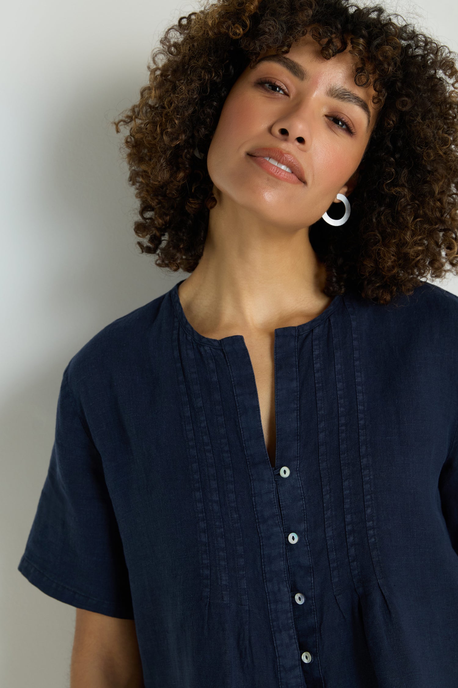 A person wears the Linen Pleat Short Sleeve Shirt, a lightweight dark blue linen top with short sleeves, round neckline, vertical front pleats, and button-up design.