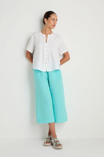 A woman stands against a white wall wearing the Linen Pleat Short Sleeve White Shirt, turquoise wide-leg pants, and silver sandals.