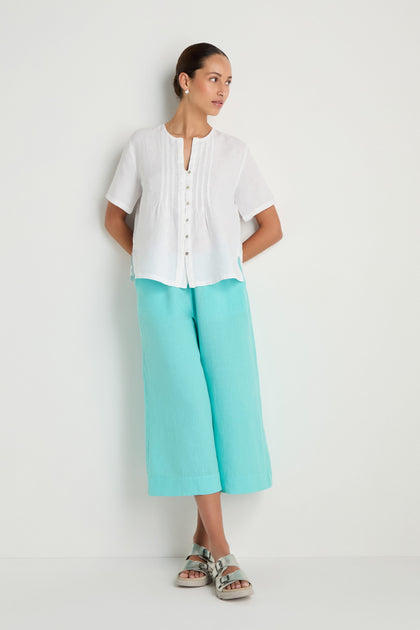 A woman stands against a white wall wearing the Linen Pleat Short Sleeve White Shirt, turquoise wide-leg pants, and silver sandals.