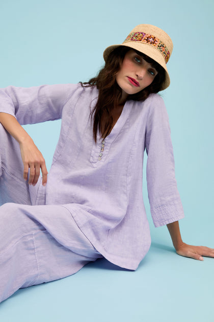 A woman wears the Linen Seam Flare Lilac Tunic and a beige hat with a patterned band while sitting on the floor against a light blue background.