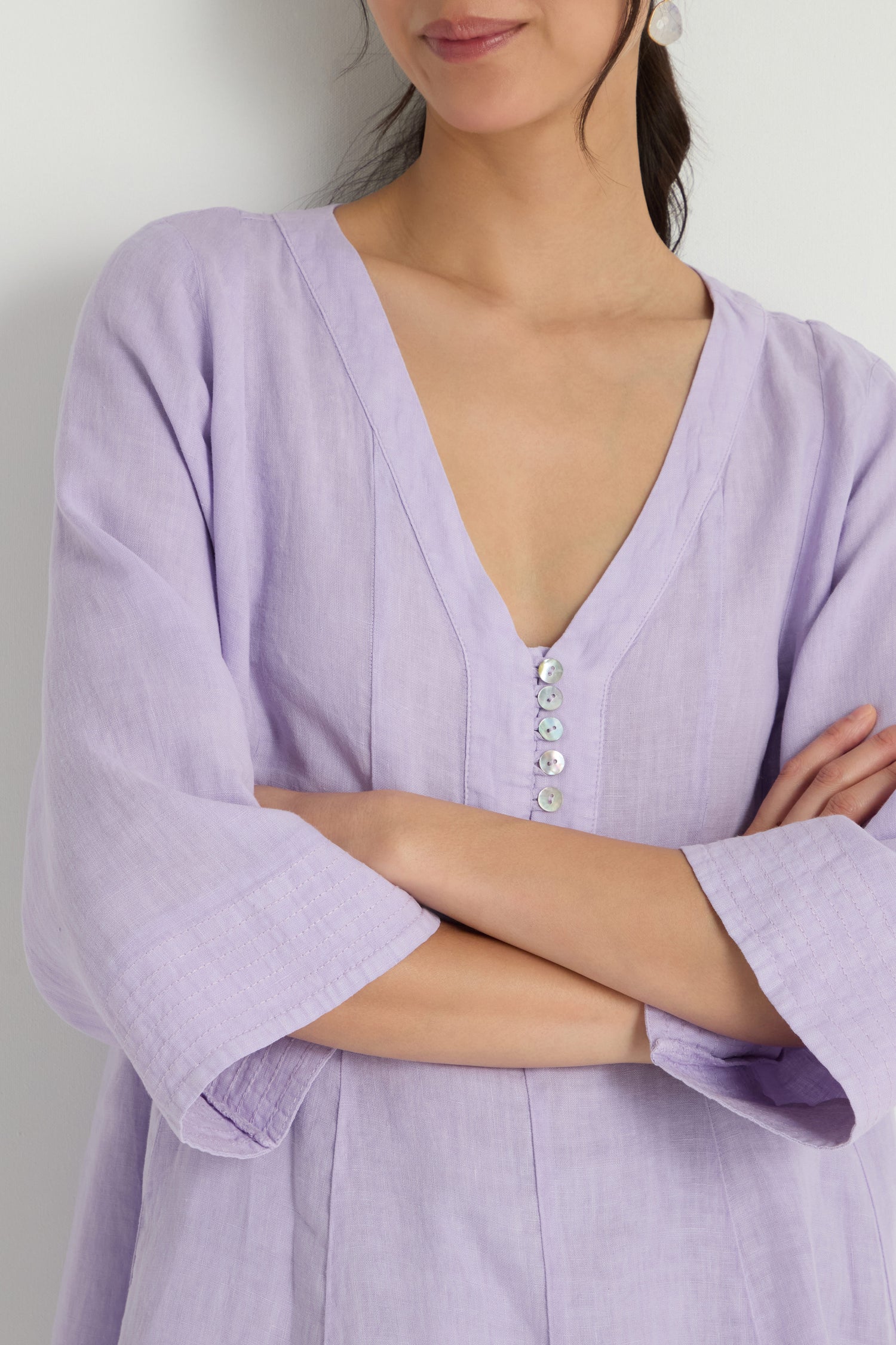 A woman stands with arms crossed, wearing the Linen Seam Flare Lilac Tunic—a light purple V-neck with three-quarter sleeves and a gentle flare silhouette. Her face is partially out of frame.
