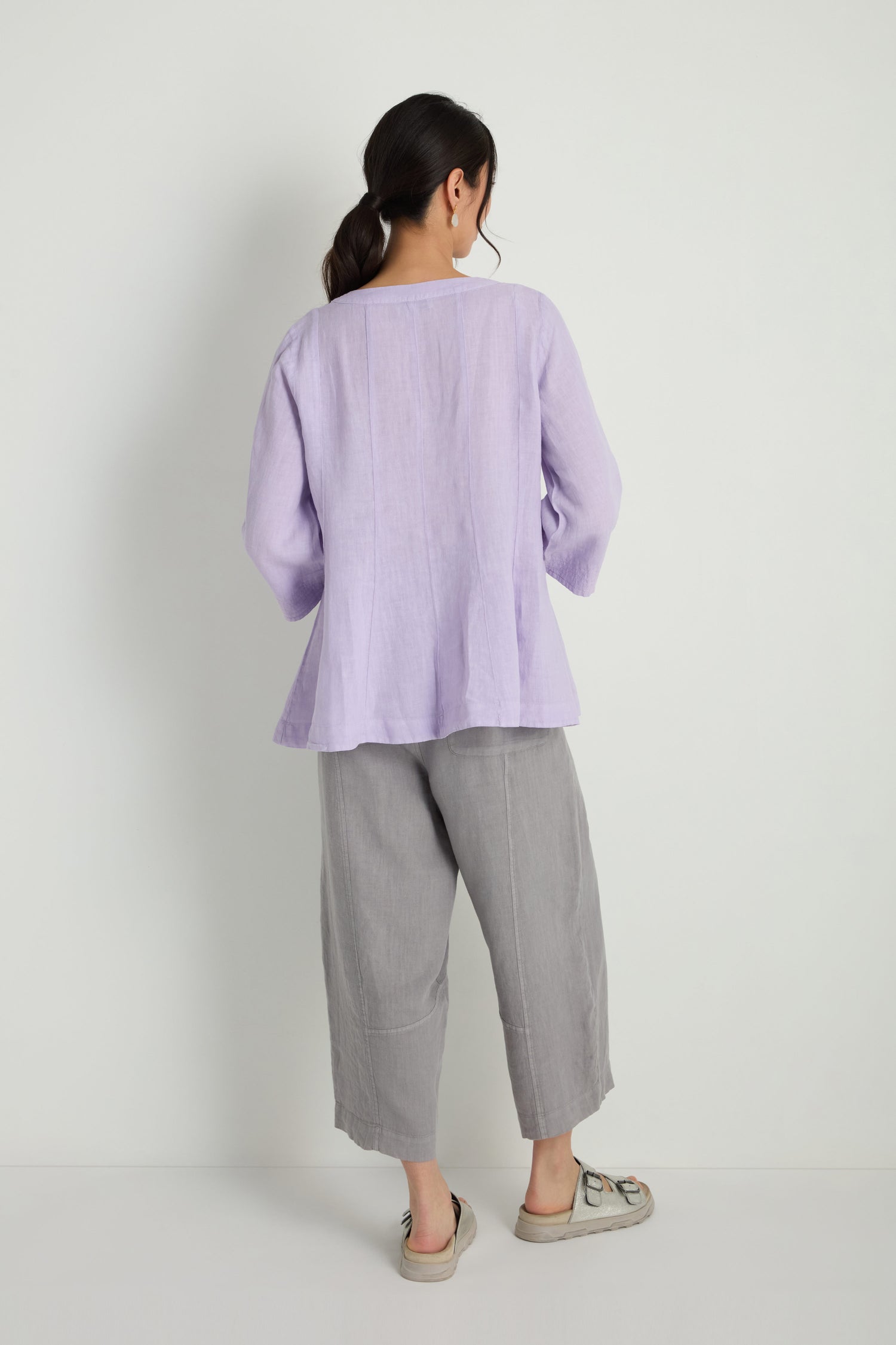 A woman stands with her back to the camera wearing the Linen Seam Flare Lilac Tunic, paired with loose gray flare pants and beige shoes, set against a plain white background.