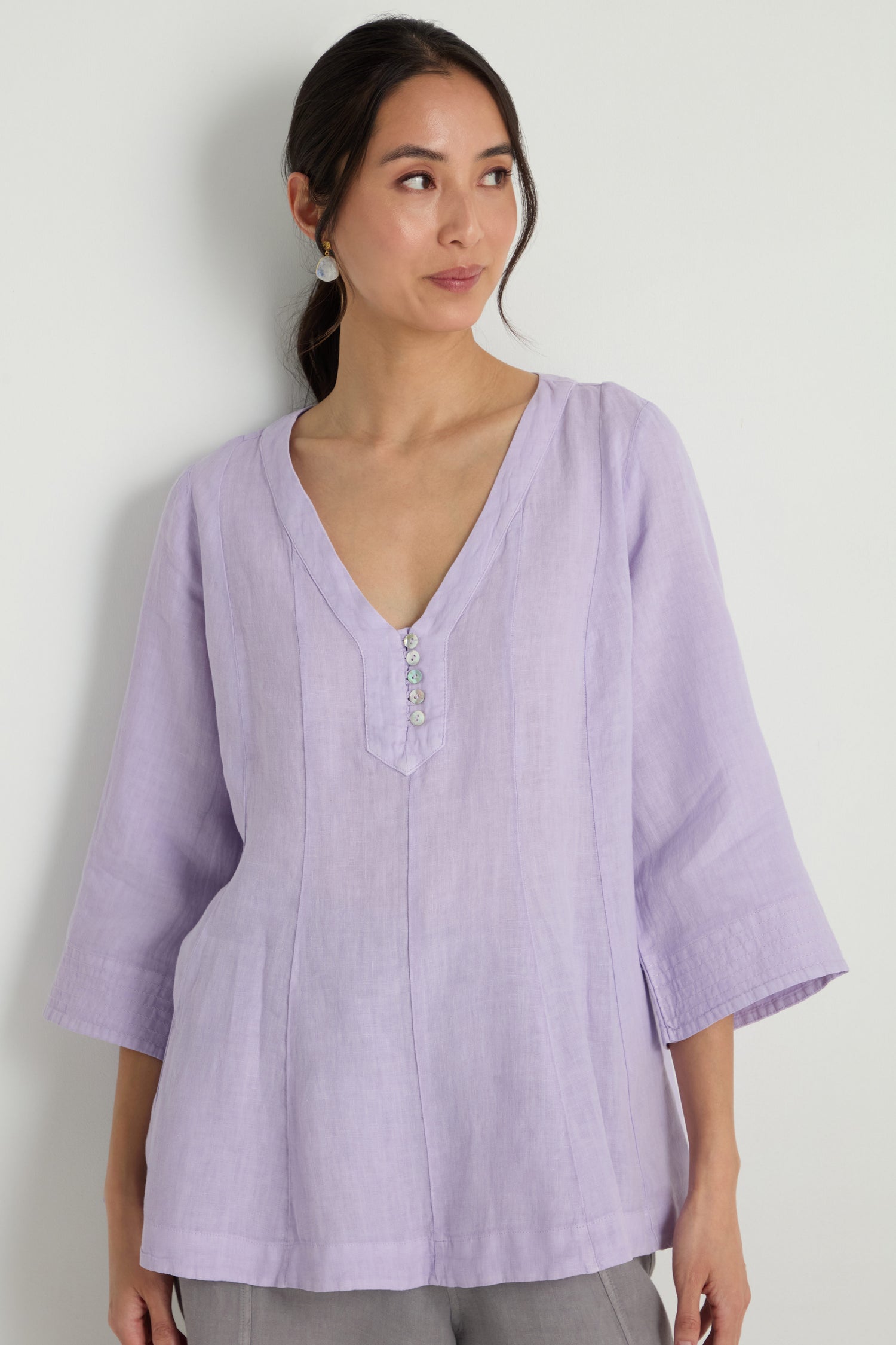 A woman stands against a plain white background wearing the Linen Seam Flare Lilac Tunic, featuring a light purple hue, V-neckline, three decorative buttons, and three-quarter subtly flared sleeves.