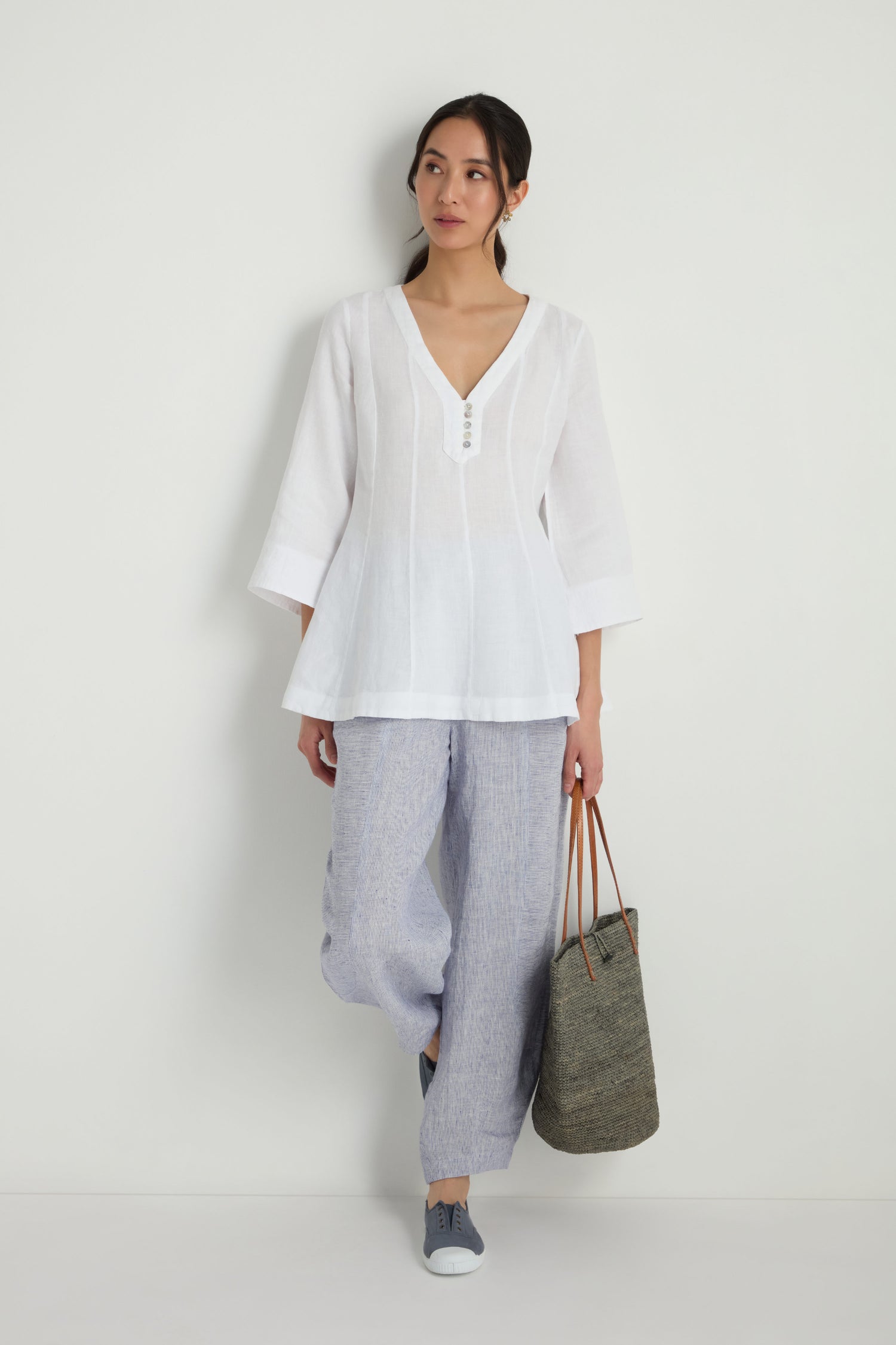 A woman stands against a white wall wearing the Linen Seam Flare White Tunic with three-quarter sleeves, paired with loose light gray pants, blue sneakers, and holding a large woven tote.