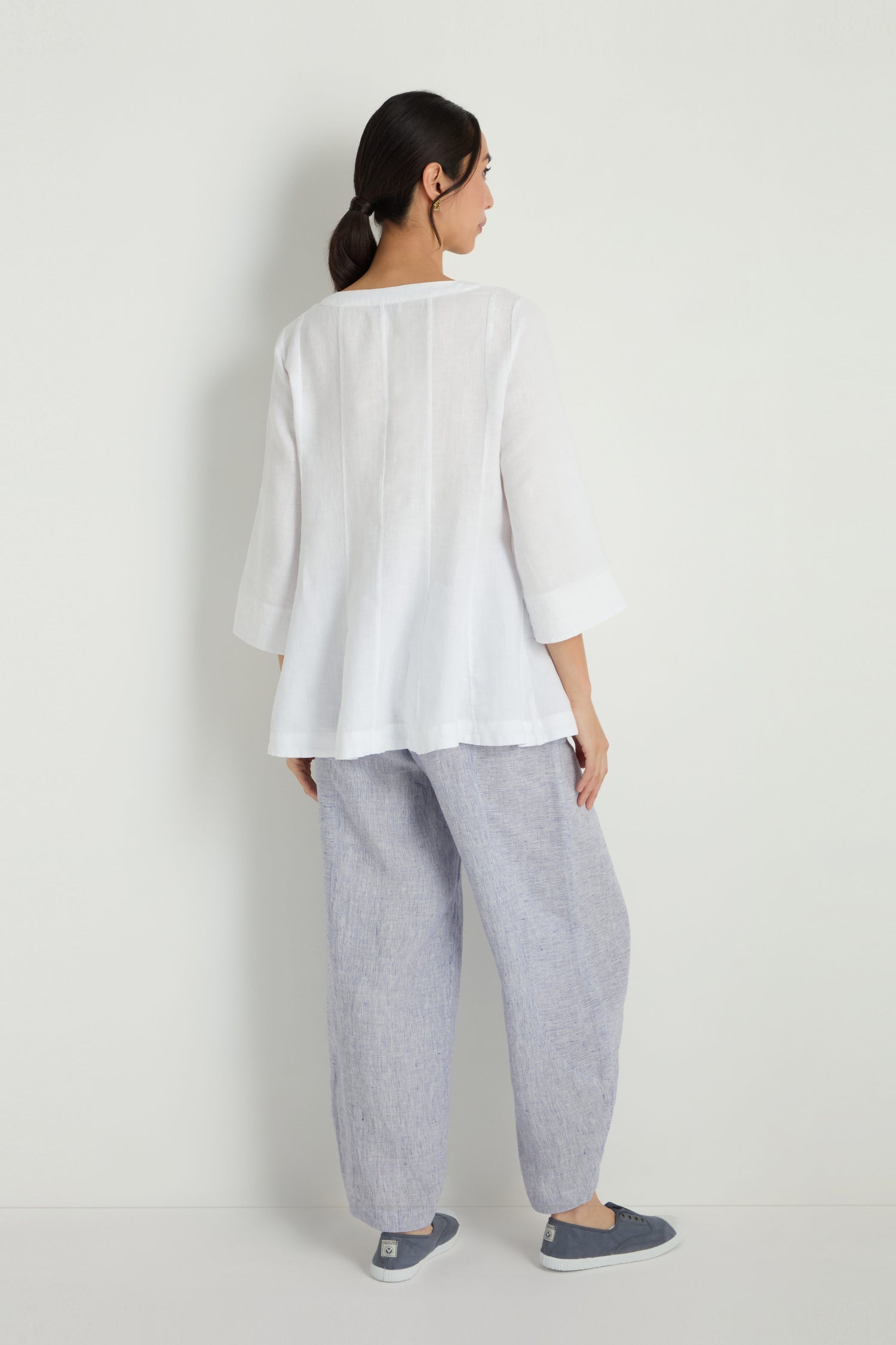 A woman stands against a plain wall, wearing the Linen Seam Flare White Tunic—loose with three-quarter sleeves—paired with light gray wide-leg pants and gray slip-on shoes, facing away with her head slightly turned.