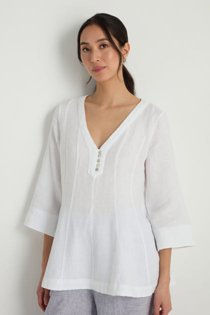 A woman wears the Linen Seam Flare White Tunic, featuring a loose fit, V-neck, button accents, and a gently flared silhouette, standing against a plain light background.