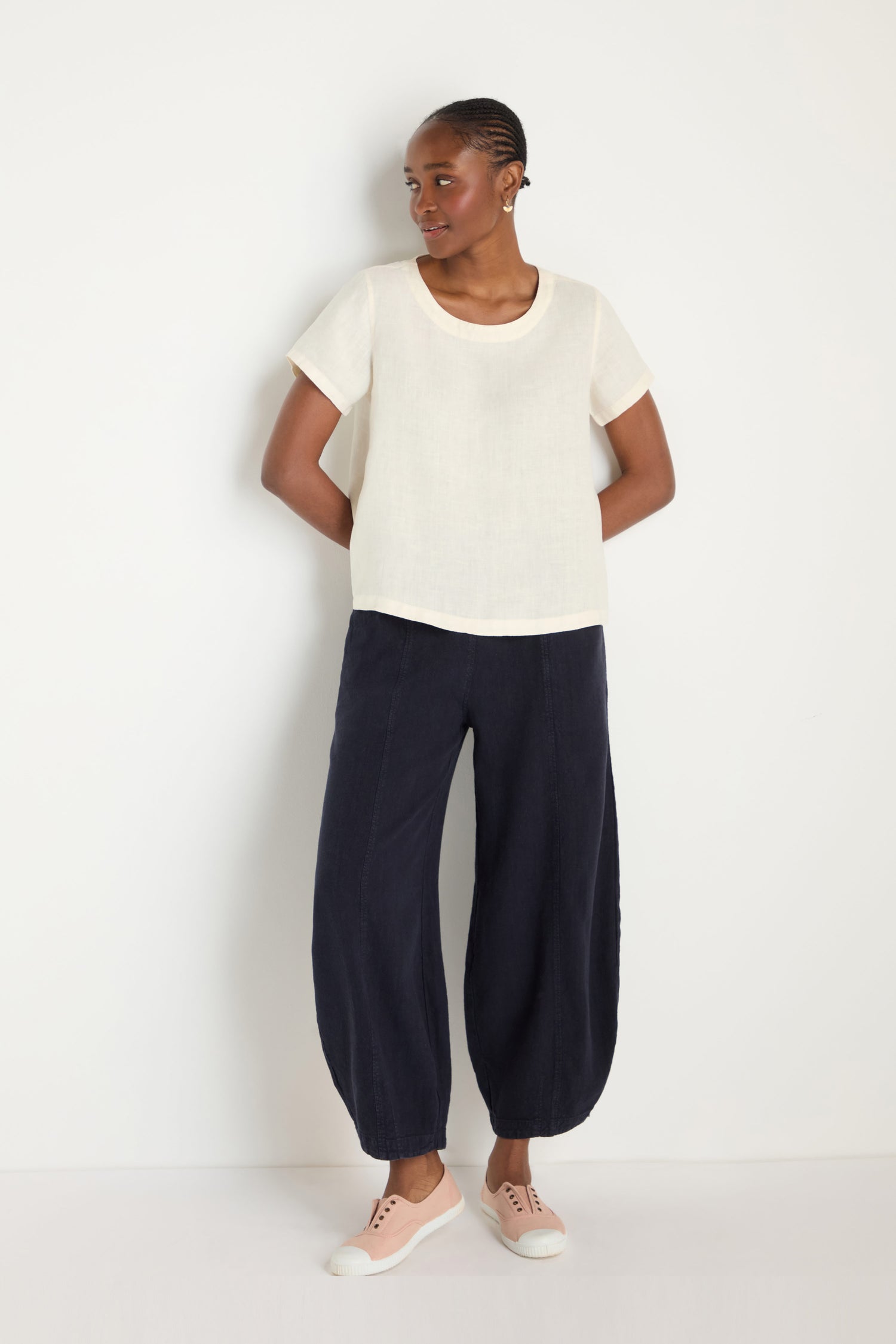 A woman stands against a white wall wearing an Ecru Linen T-shirt, loose dark blue pants, and light pink sneakers.
