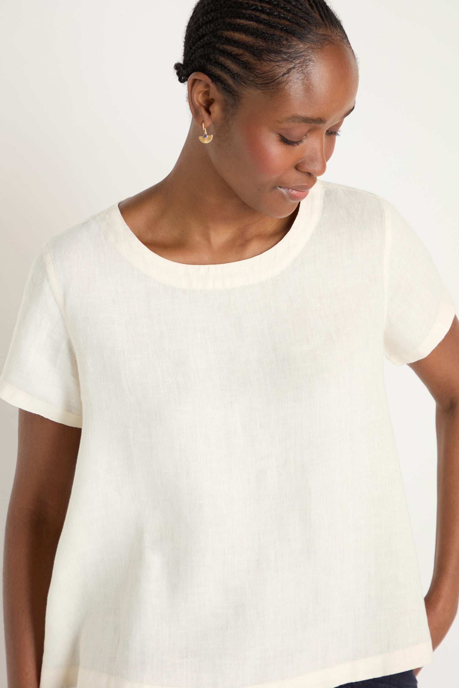 A person wearing the Ecru Linen T-shirt, a simple short-sleeve pure linen top, looks down slightly with one hand on their hip against a plain background.
