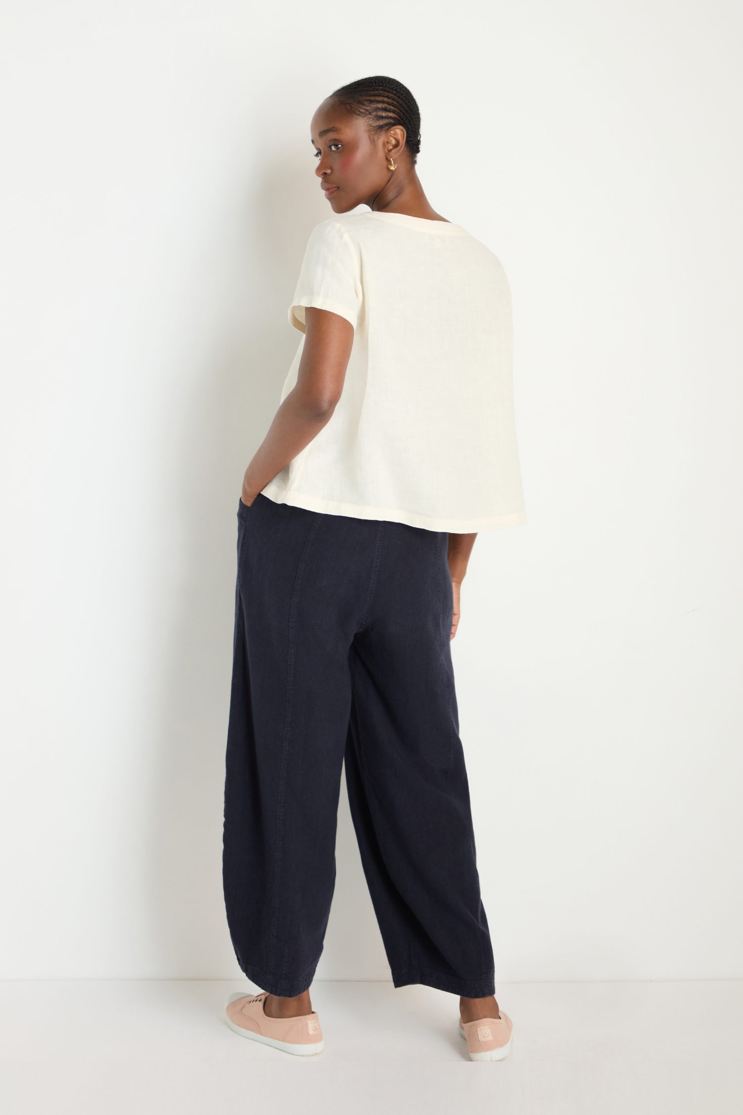 A person stands against a white wall, glancing back over their shoulder. They wear an Ecru Linen T-shirt with short sleeves, paired with wide-leg dark pants and light slip-on shoes.