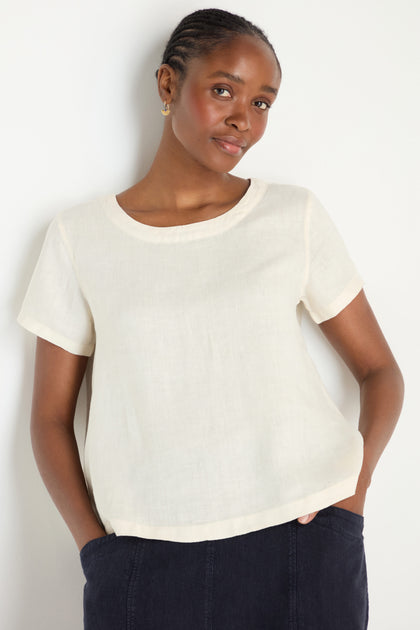 A woman stands against a plain white background with her hands in her pockets, wearing an Ecru Linen T-shirt and dark pants.