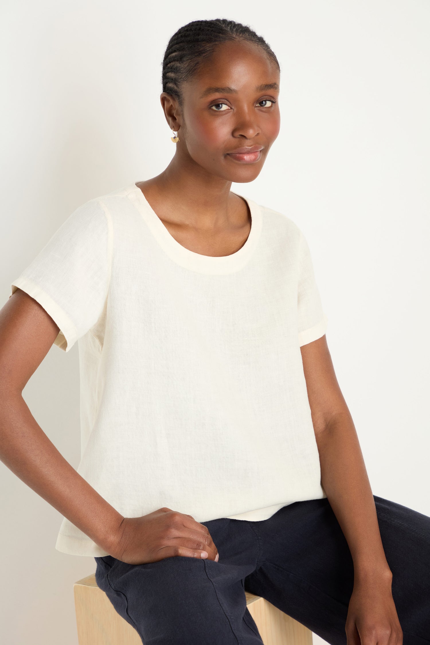 A woman with short, braided hair sits on a light stool, wearing an Ecru Linen T-shirt and dark pants against a plain light background.