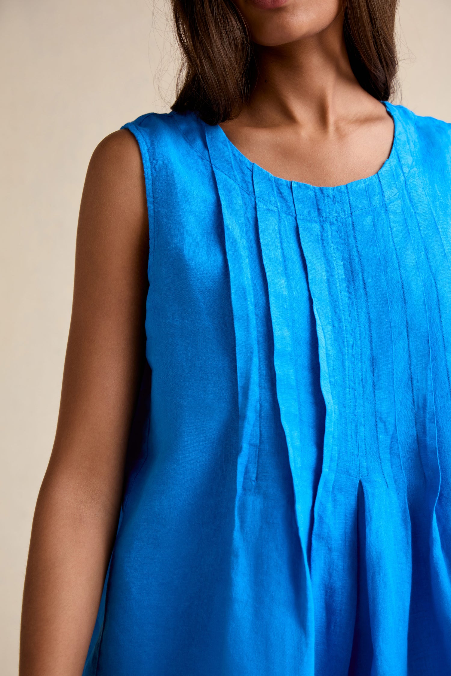 A woman models the Linen Pleat Front Azure Vest, a sleeveless blue linen top with pleated detailing, shown from shoulders to mid-torso against a neutral background.