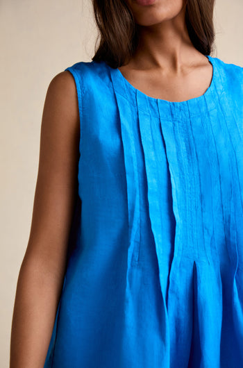 A woman models the Linen Pleat Front Azure Vest, a sleeveless blue linen top with pleated detailing, shown from shoulders to mid-torso against a neutral background.