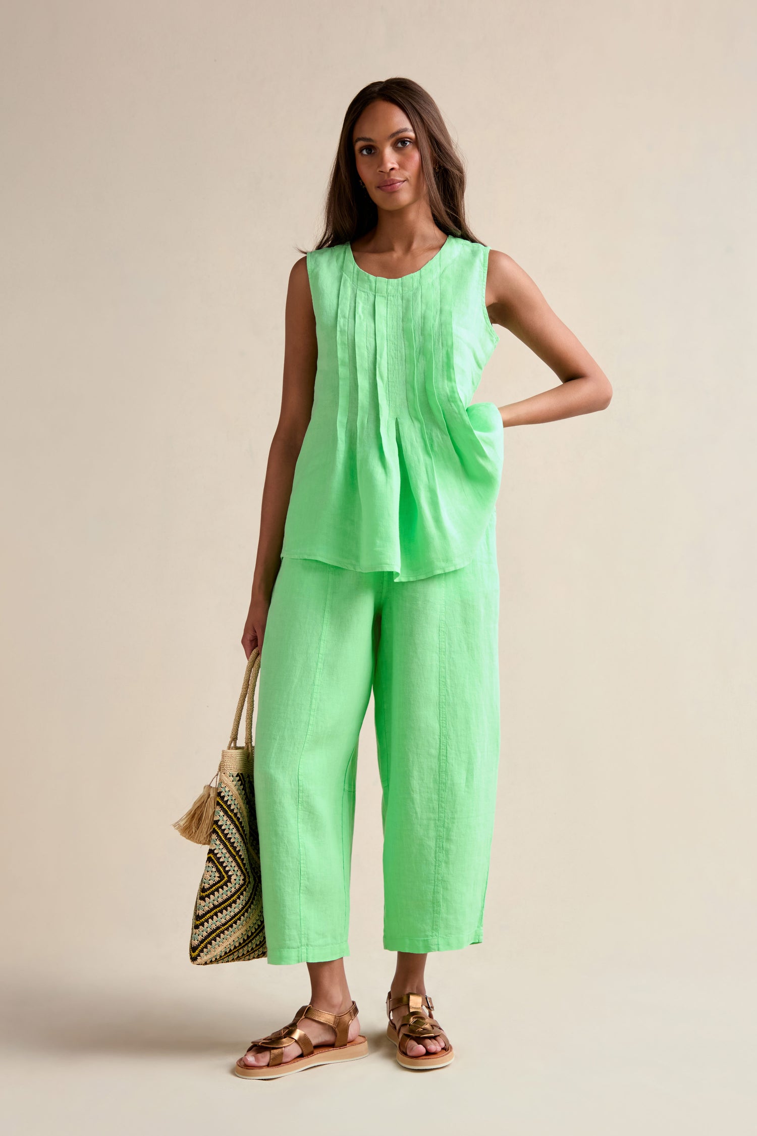 A woman stands against a neutral background wearing the Linen Pleat Front Pistachio Vest with matching wide-leg pants, holding a woven tote bag and wearing brown sandals.