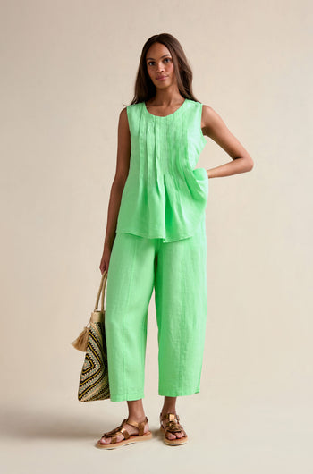 A woman stands against a neutral background wearing the Linen Pleat Front Pistachio Vest with matching wide-leg pants, holding a woven tote bag and wearing brown sandals.