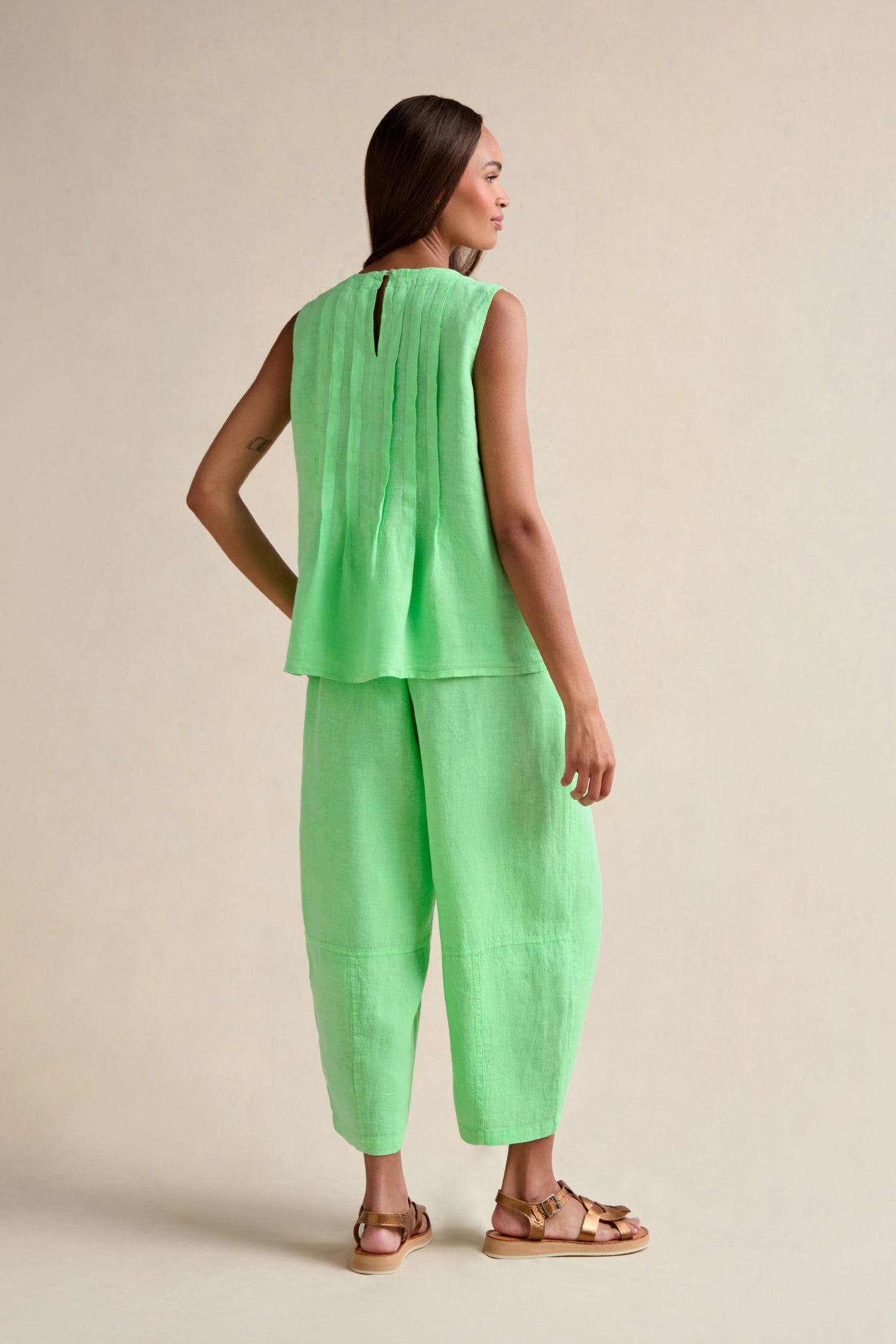 A woman faces backward, wearing the Linen Pleat Front Pistachio Vest with matching loose pants and flat brown sandals.