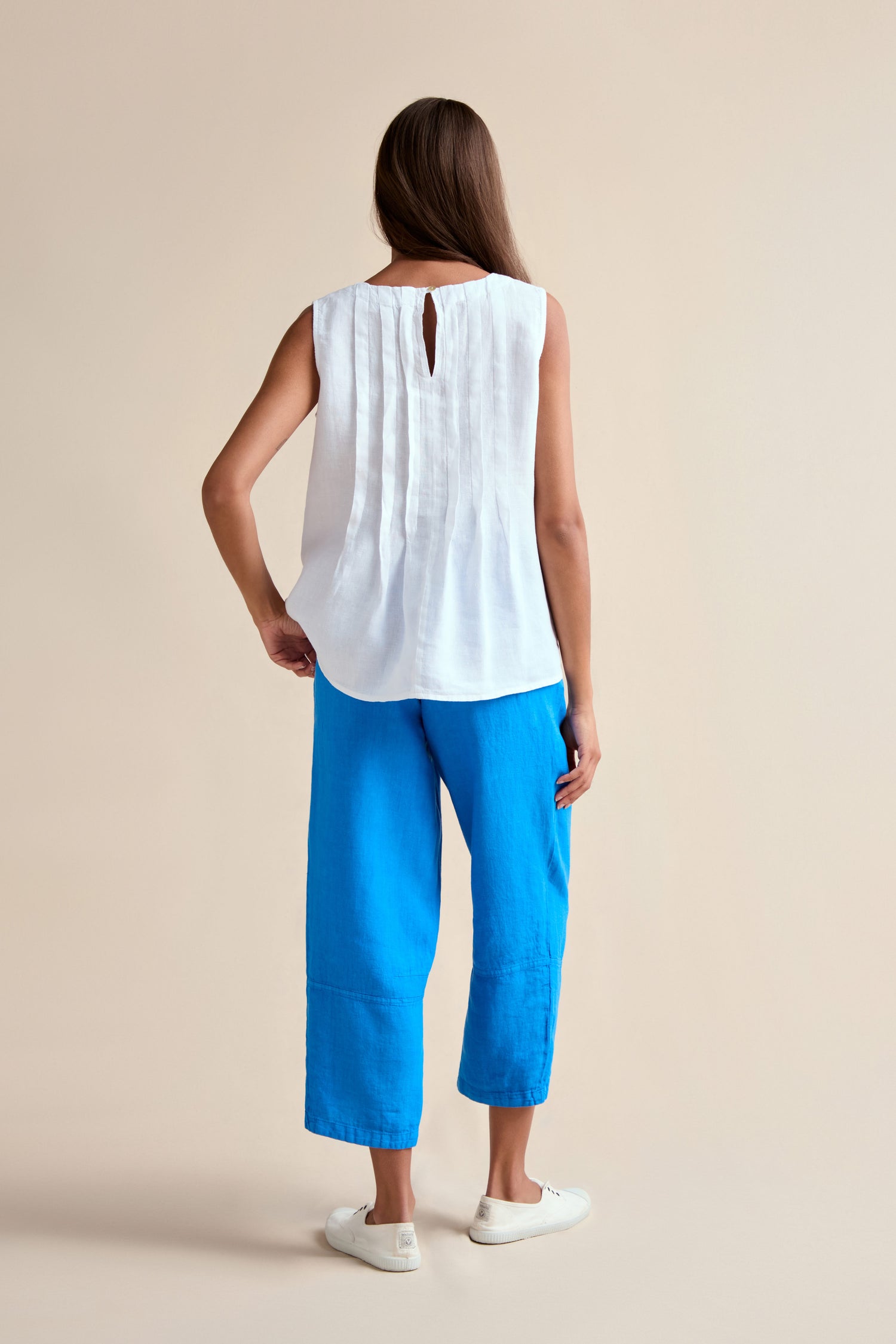 A woman faces away, wearing the Linen Pleat Front White Vest, bright blue cropped pants, and white slip-on shoes against a plain beige background.