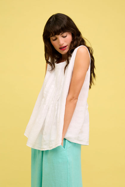 A woman with long brown hair wears the Linen Pleat Front White Vest and light blue pants, standing against a plain yellow background, looking down with her hands in her pockets.