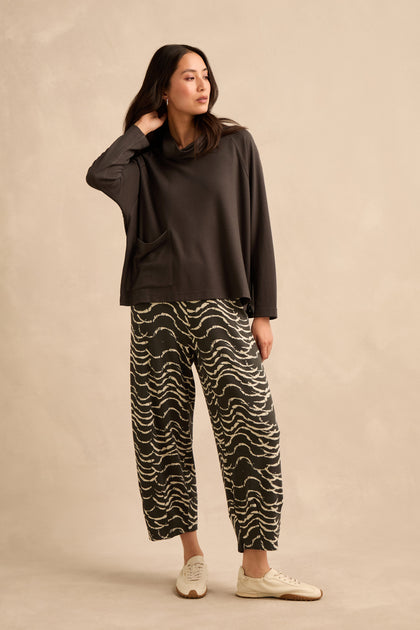 A woman stands against a beige background, wearing the Luxe Viscose Jersey Cowl Neck Top, patterned wide-leg pants, and white sneakers, with one hand touching her neck.