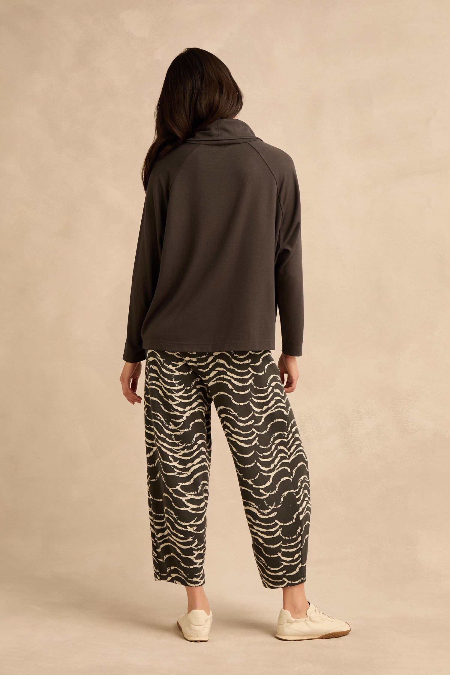 Person standing against a beige background, facing away, wearing the Luxe Viscose Jersey Cowl Neck Top, patterned wide-leg pants, and light-colored shoes.