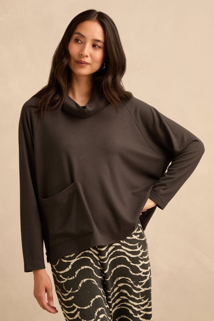 A woman wearing a Luxe Viscose Jersey Cowl Neck Top with long sleeves and a front pocket, paired with patterned black and white pants, stands against a beige background.