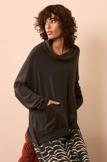 A woman with curly hair wears the Luxe Viscose Jersey Cowl Neck Top in black, styled with patterned pants, standing against a neutral background with one hand in her pocket.