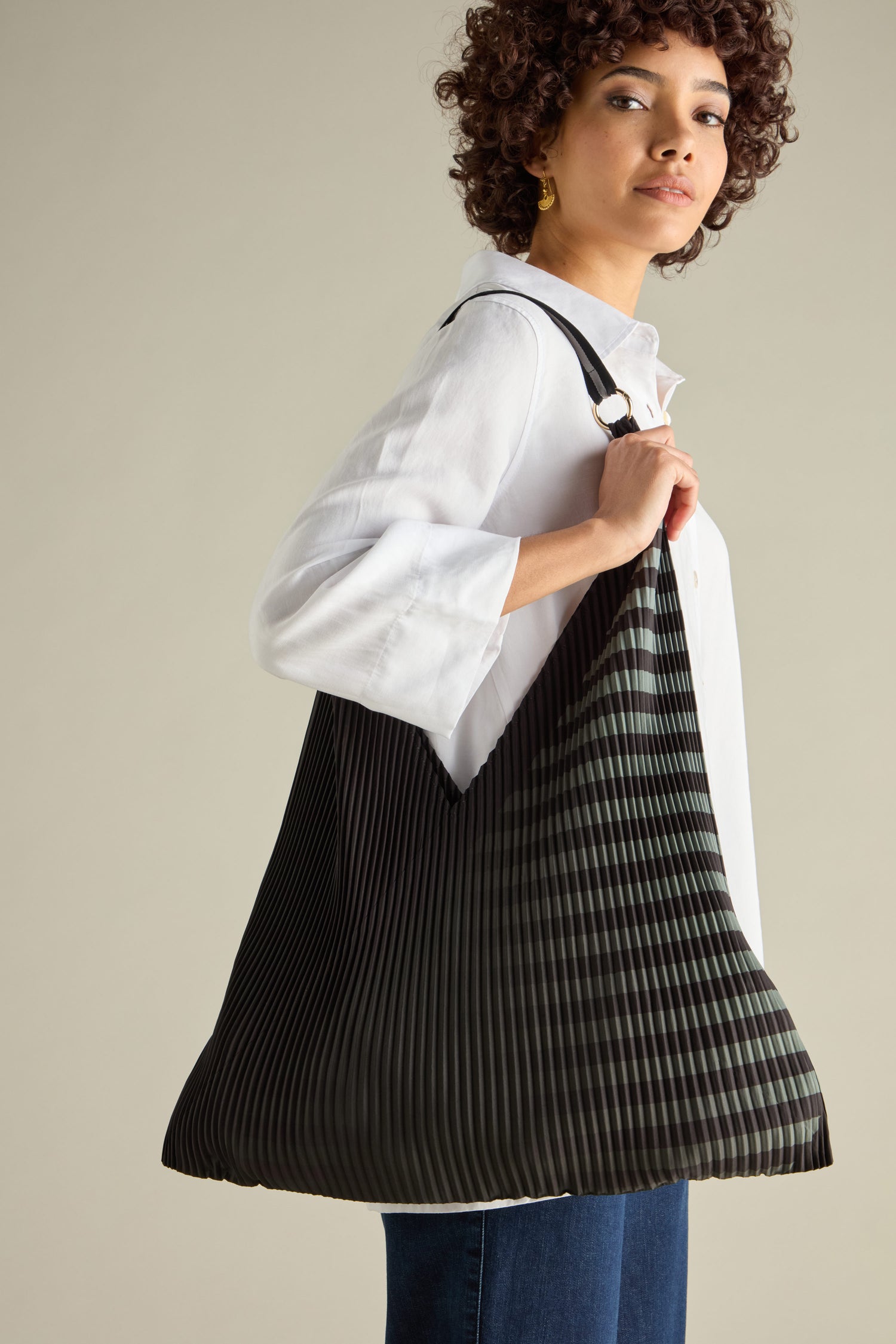 A person with curly hair, in a white shirt and blue jeans, casually carrying a large eco-friendly Pleated Y Handbag with black and green stripes.