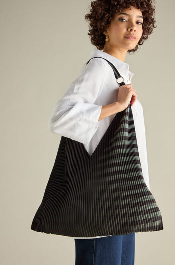 A person with curly hair, in a white shirt and blue jeans, casually carrying a large eco-friendly Pleated Y Handbag with black and green stripes.