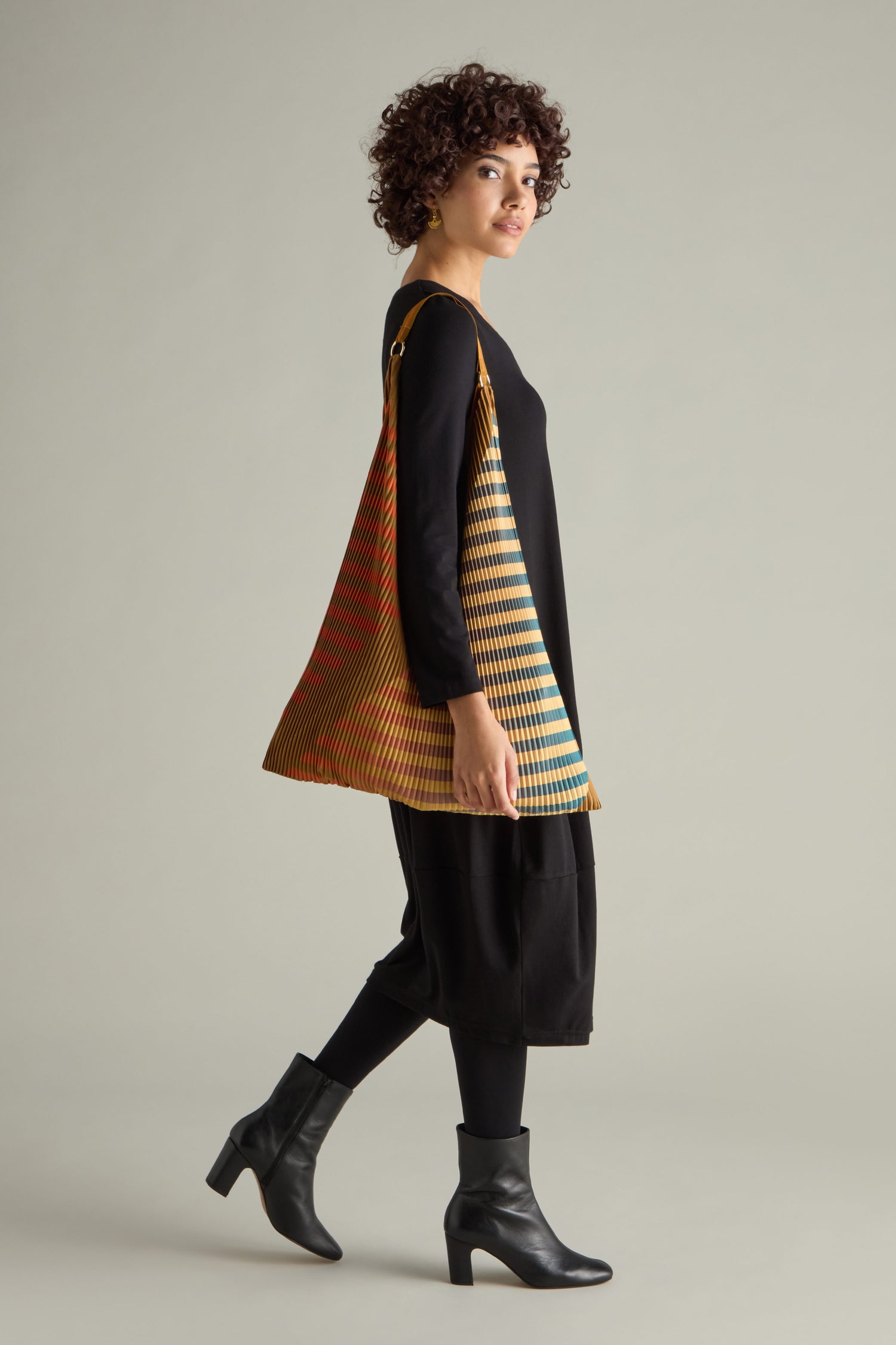 A model with curly hair showcases a black dress accented by a colorful, striped triangular accessory and black ankle boots, paired with the elegant Pleated Y Handbag, in an eco-friendly design against a plain backdrop.