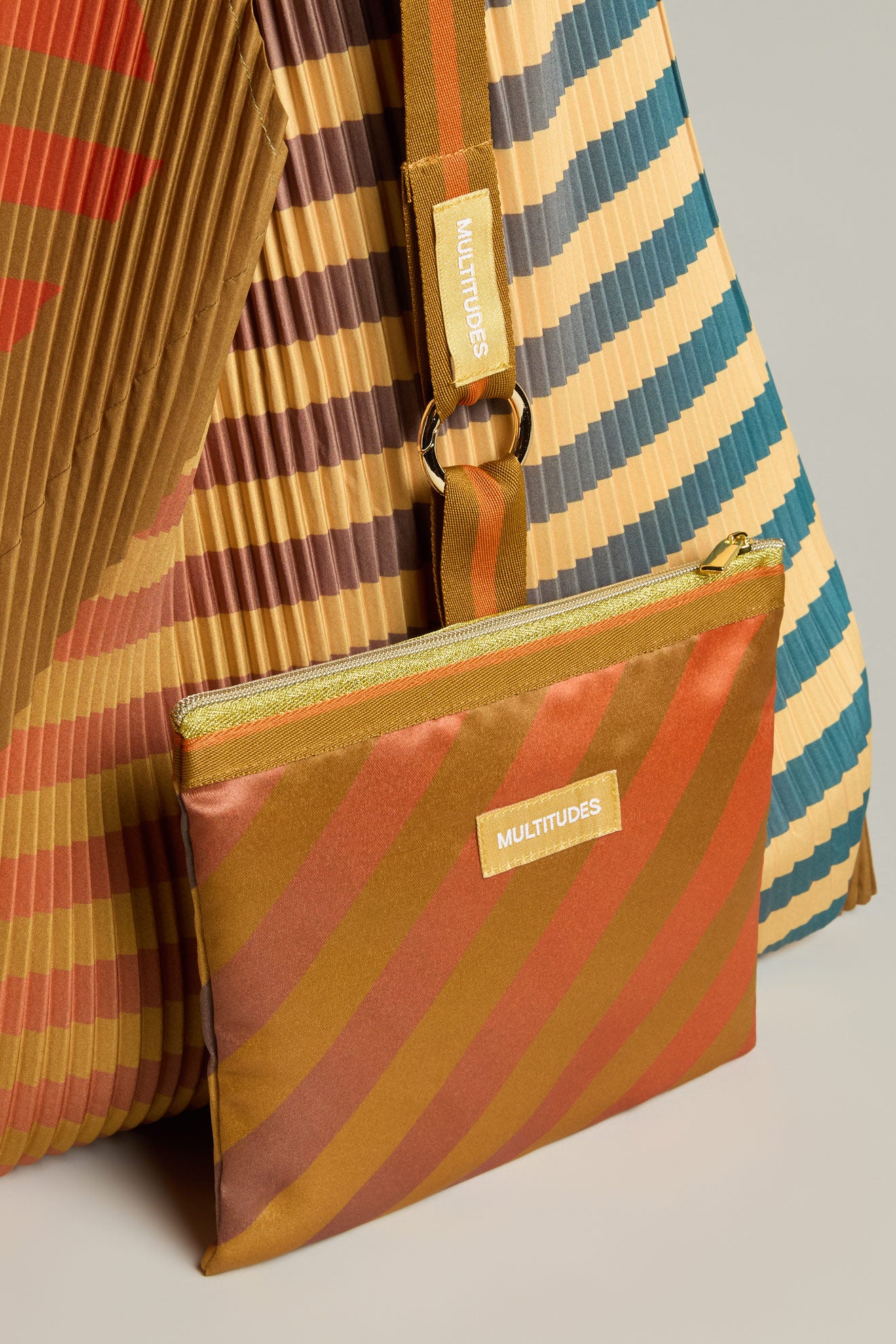 The Pleated Y Handbag, designed sustainably with vibrant stripes, includes a matching detachable pouch, both featuring the "MULTITUDES" label.