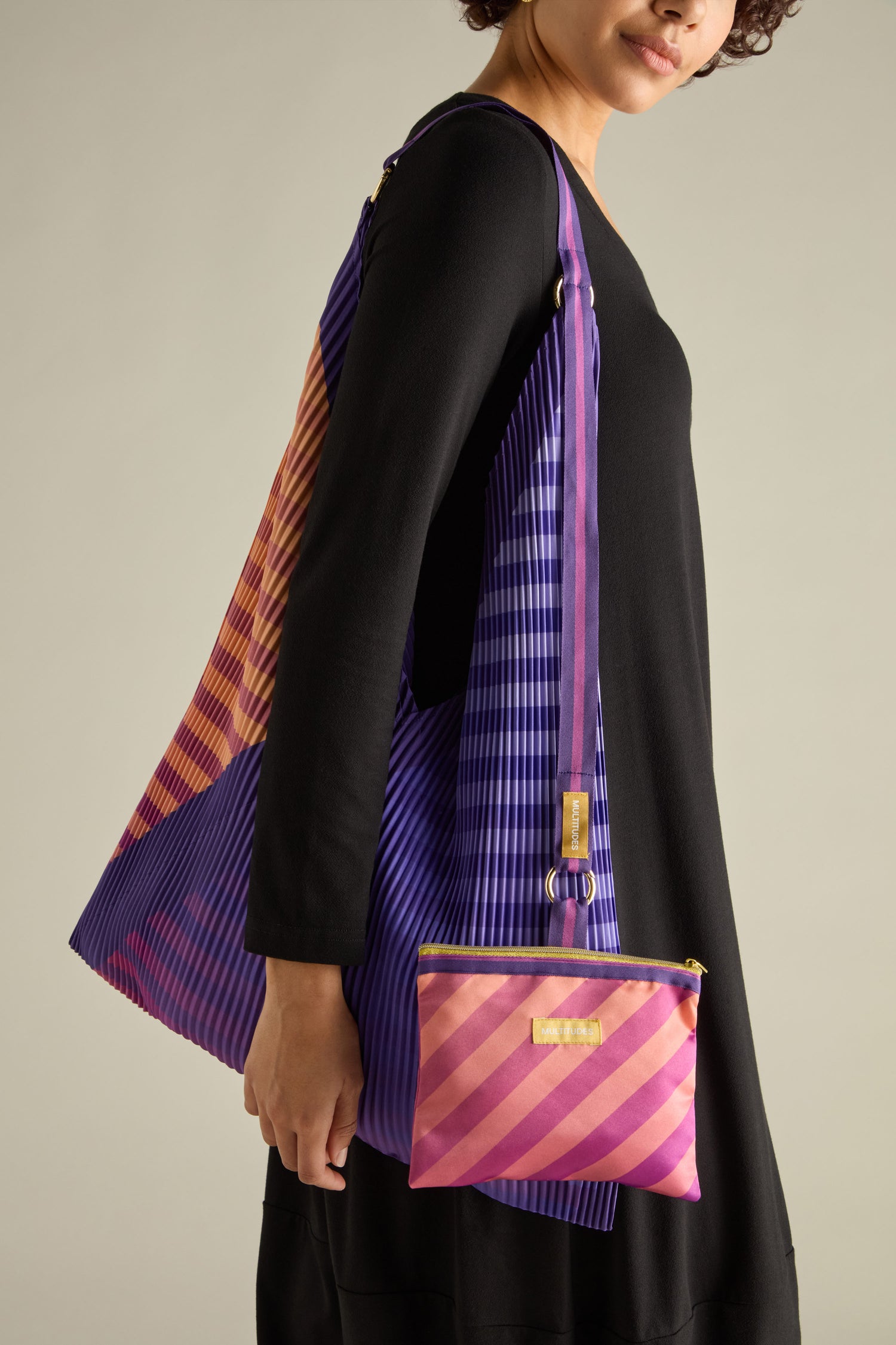 A person in a black dress holds a Pleated Y Handbag, featuring eco-friendly purple and pink stripes, along with a matching small pouch.