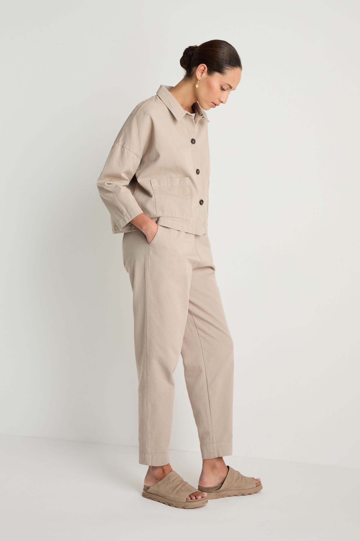 A woman stands sideways against a white background, wearing a beige Heavy Cotton Boxy Jacket, matching pants, and tan slide sandals.