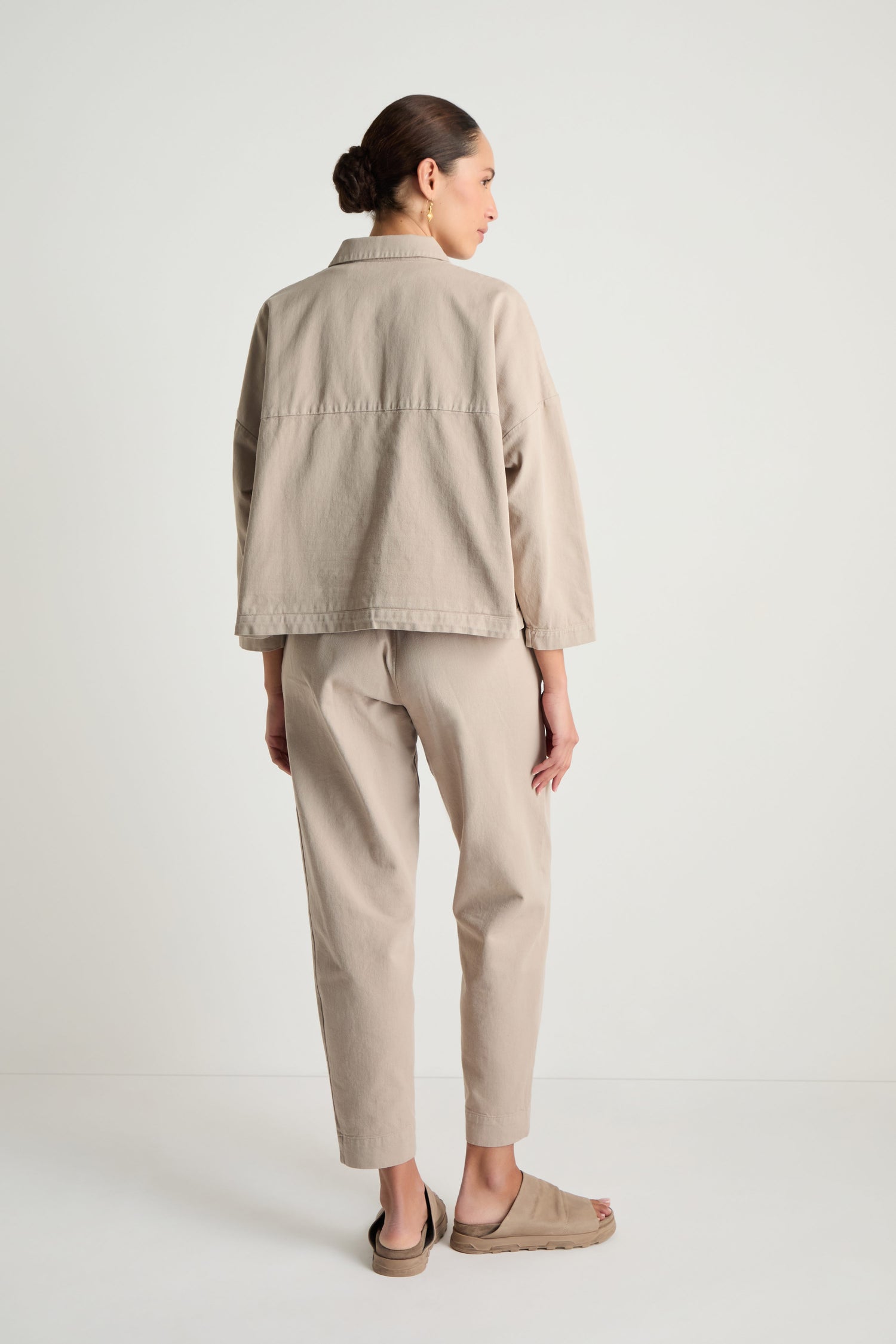 A woman faces away, wearing the Heavy Cotton Boxy Jacket in beige, with matching pants and tan sandals, set against a plain white background.