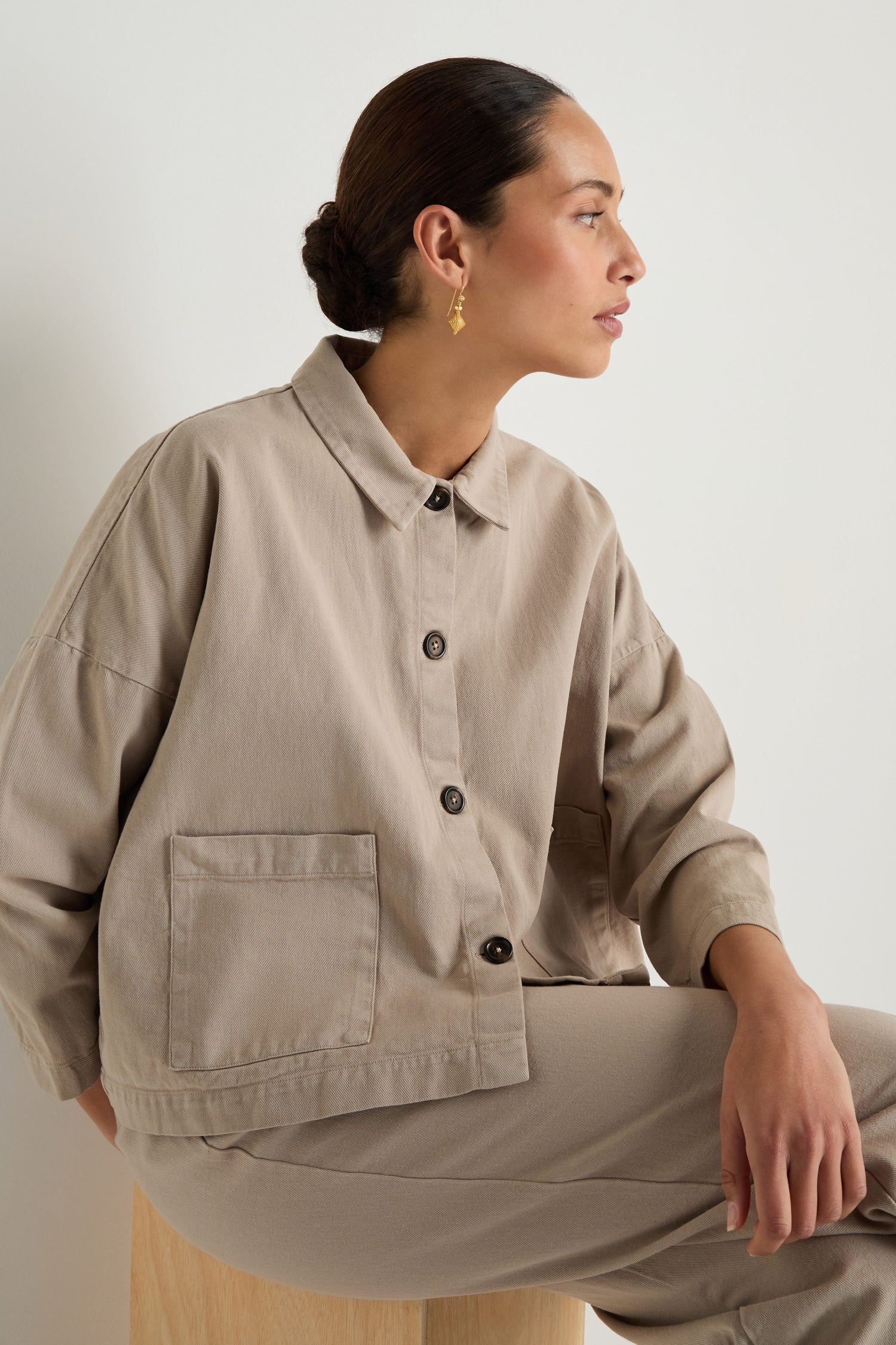A woman with dark hair in a low bun wears the Heavy Cotton Boxy Jacket and matching pants, seated on a wooden stool and looking to the side against a plain background.