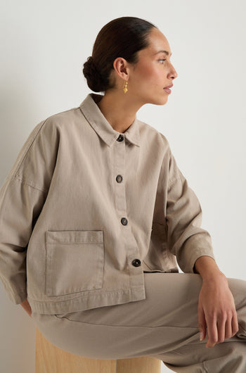 A woman with dark hair in a low bun wears the Heavy Cotton Boxy Jacket and matching pants, seated on a wooden stool and looking to the side against a plain background.