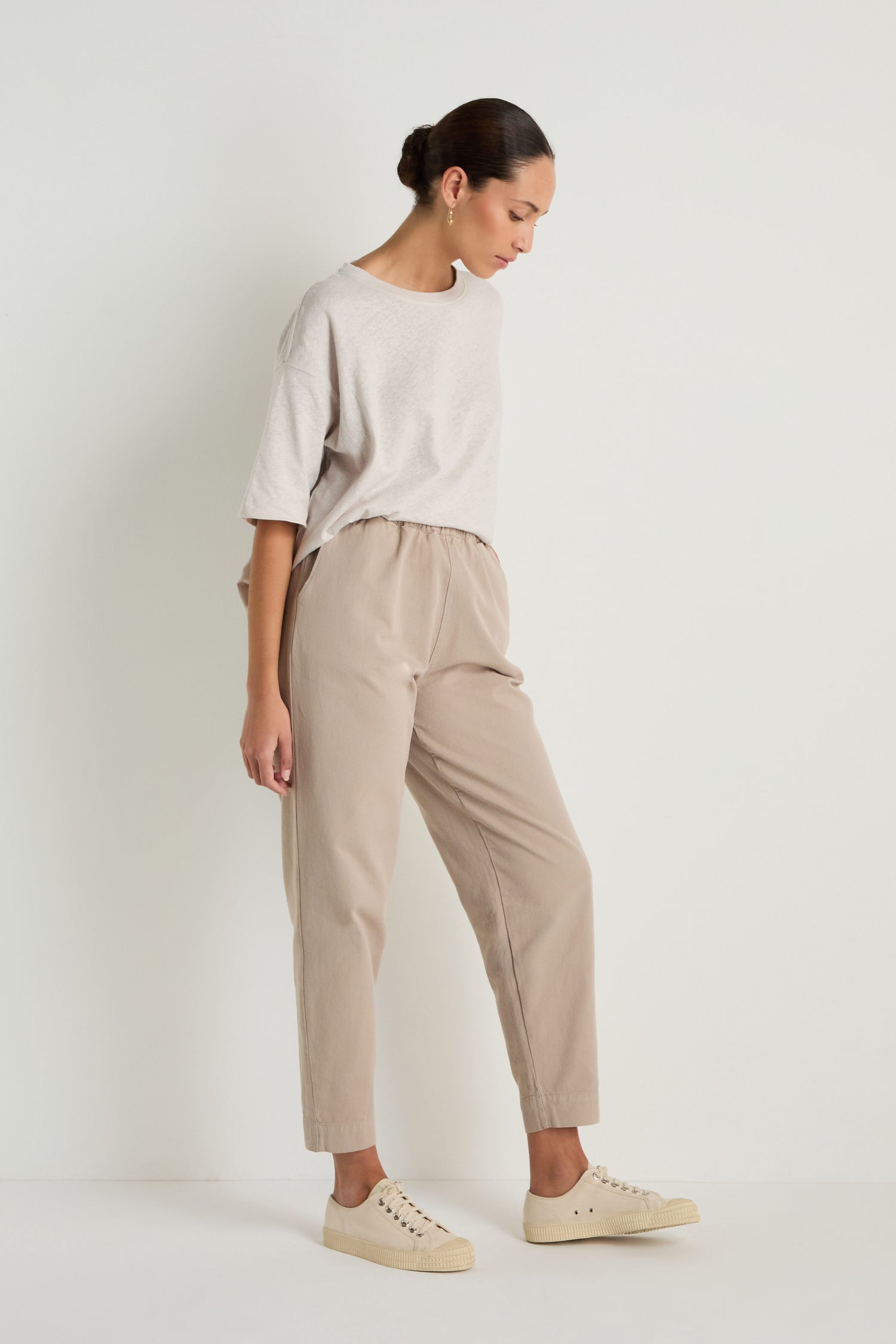 A person stands by a plain white wall, wearing loose light beige t-shirt, Heavy Cotton Straight Trousers with an elastic waist, cream sneakers, and has their hair pulled back in a neat bun.