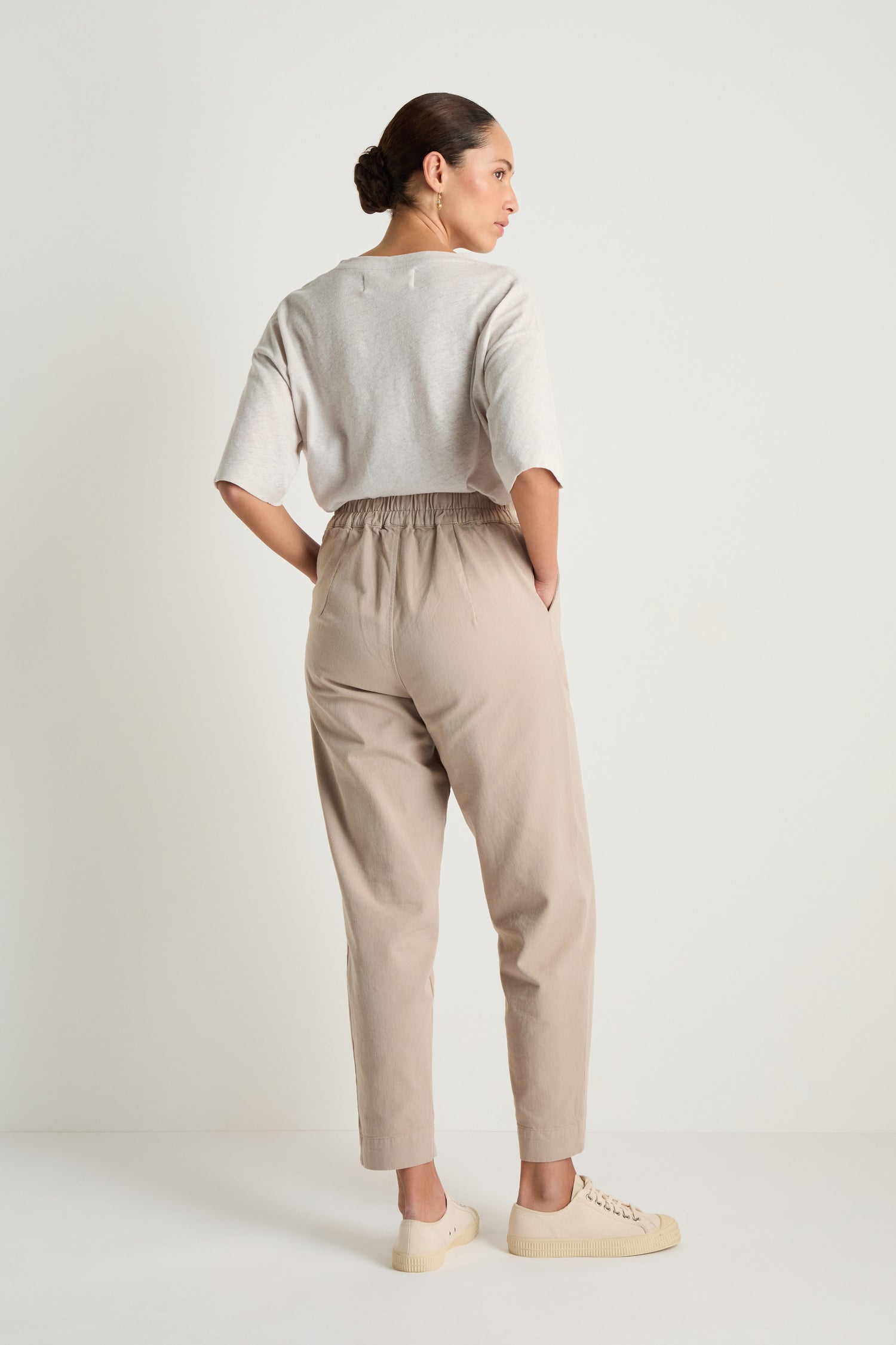 A woman faces away against a plain background wearing a light gray top and beige Heavy Cotton Straight Trousers with an elastic waist, styled with cream sneakers.