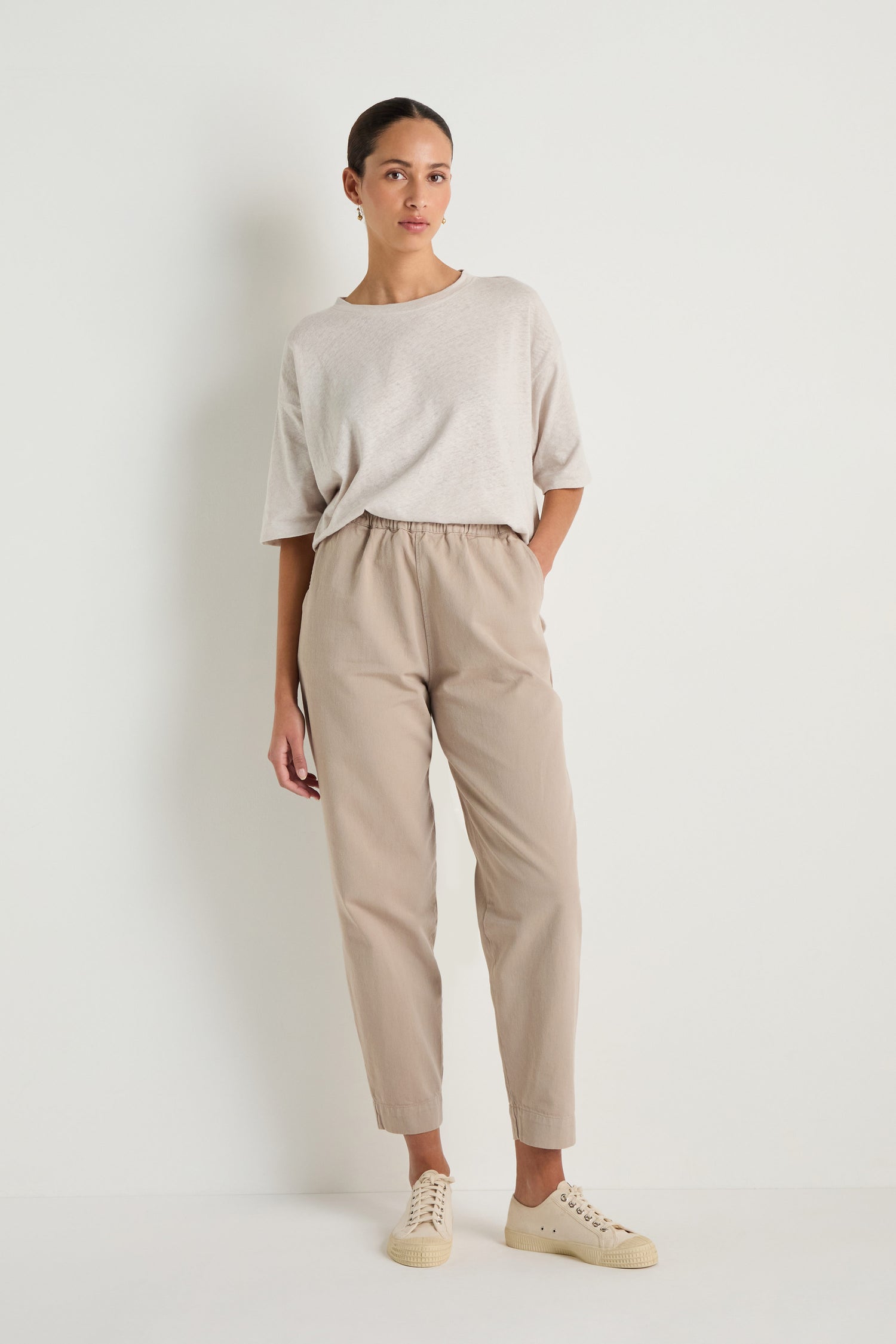 A woman stands against a plain white wall wearing a loose light beige T-shirt, Heavy Cotton Straight Trousers with an elasticated waist, and light-colored sneakers, with one hand in her pocket.