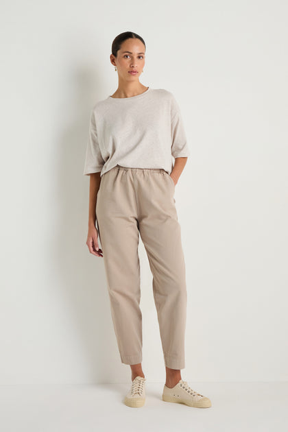 A woman stands against a plain white wall wearing a loose light beige T-shirt, Heavy Cotton Straight Trousers with an elasticated waist, and light-colored sneakers, with one hand in her pocket.