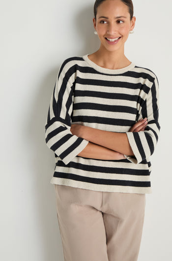 Smiling with arms crossed against a white wall, the person wears the Striped Cotton Knit sweater and beige pants—a versatile, breathable layer perfect for any season.