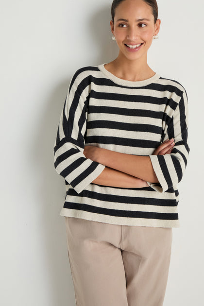 Smiling with arms crossed against a white wall, the person wears the Striped Cotton Knit sweater and beige pants—a versatile, breathable layer perfect for any season.