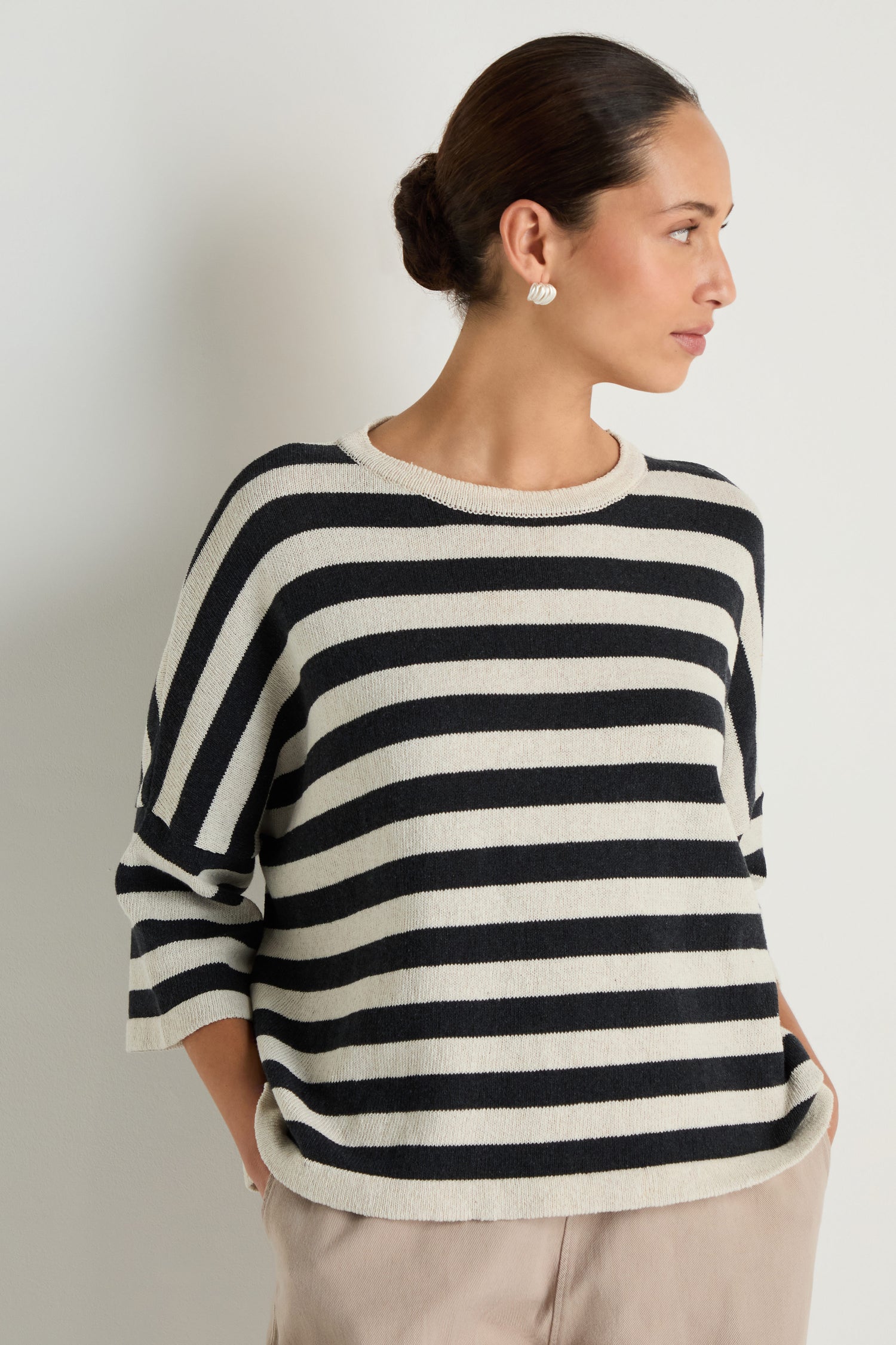 A woman models the Striped Cotton Knit, an oversized sweater, paired with beige pants as she stands with her hands in her pockets against a plain background.