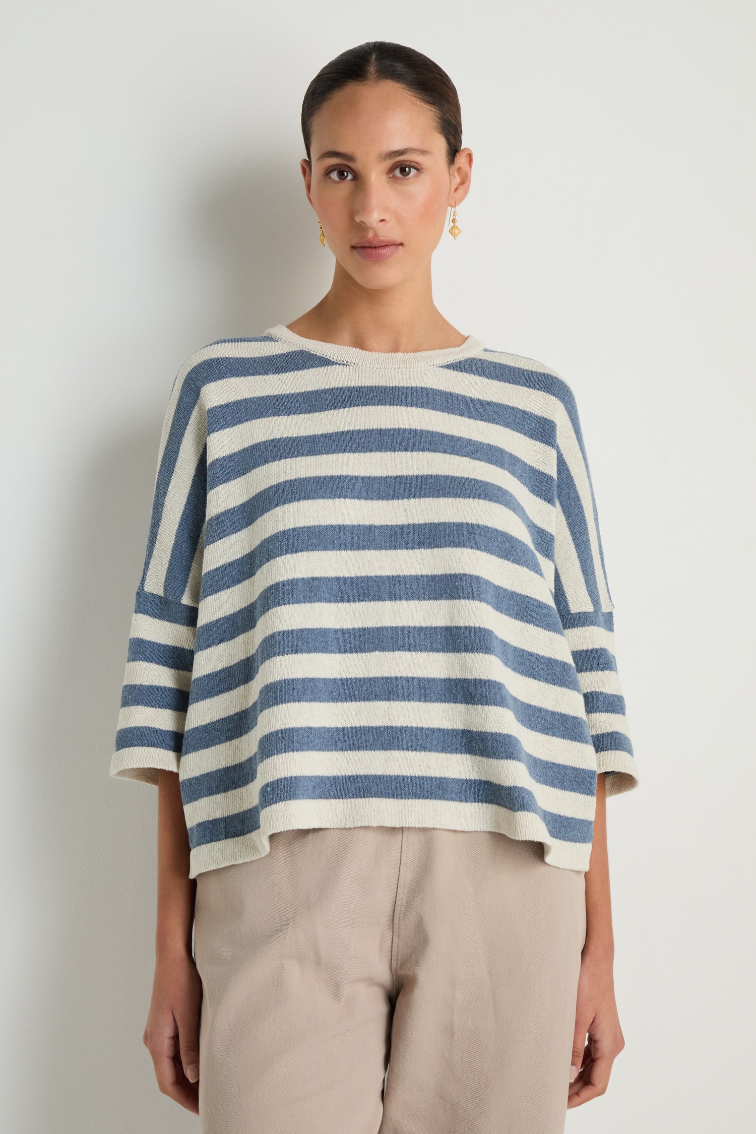 A person stands against a plain background wearing the Striped Cotton Knit, an oversized blue and white sweater, paired with beige pants for a stylish and breathable look.