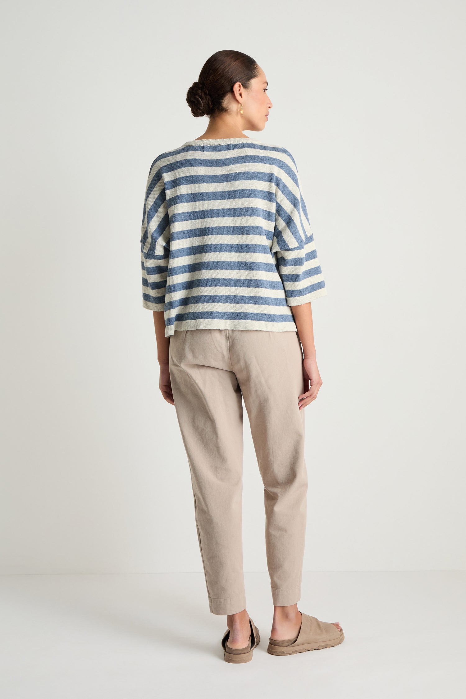 A person stands facing away, wearing the Striped Cotton Knit top in blue and white, with beige pants and tan slip-on shoes against a plain white background.